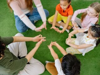 Group of children and adults sitting in a circle on the grass, doing a hand exercise