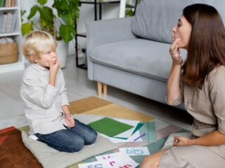 A child with blond hair sitting on a cushion and mimicking speech sounds with a female therapist, highlighting a session focused on addressing voice disorders and speech therapy.