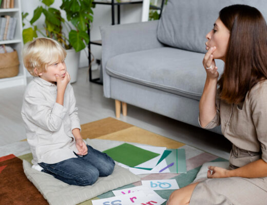 A child with blond hair sitting on a cushion and mimicking speech sounds with a female therapist, highlighting a session focused on addressing voice disorders and speech therapy.