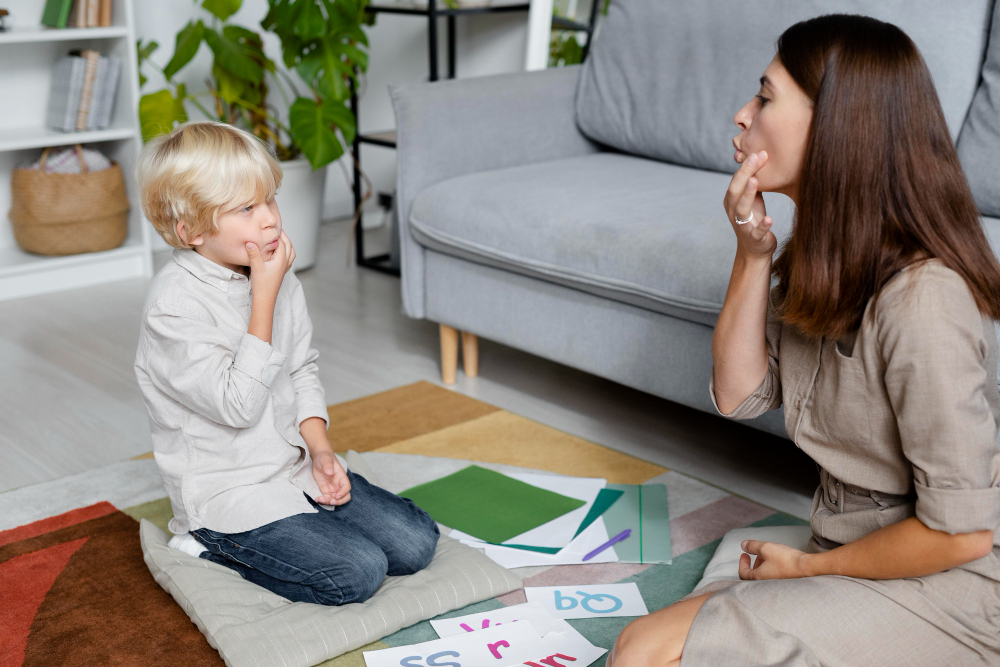 A child with blond hair sitting on a cushion and mimicking speech sounds with a female therapist, highlighting a session focused on addressing voice disorders and speech therapy.