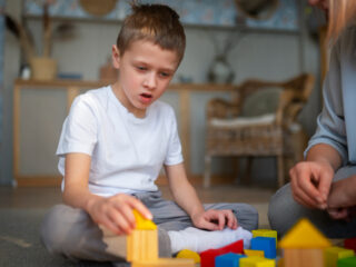 A young boy with autism sits on the floor, focused on playing with colorful building blocks. A caregiver or therapist sits nearby, assisting him.