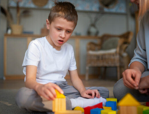 A young boy with autism sits on the floor, focused on playing with colorful building blocks. A caregiver or therapist sits nearby, assisting him.