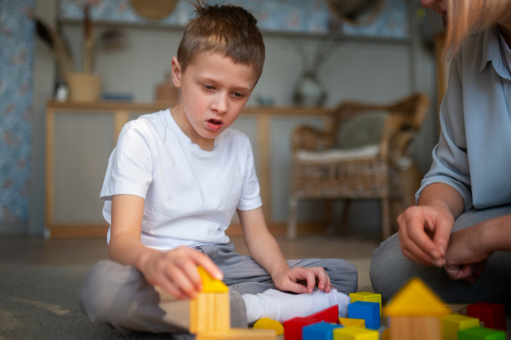 A young boy with autism sits on the floor, focused on playing with colorful building blocks. A caregiver or therapist sits nearby, assisting him.